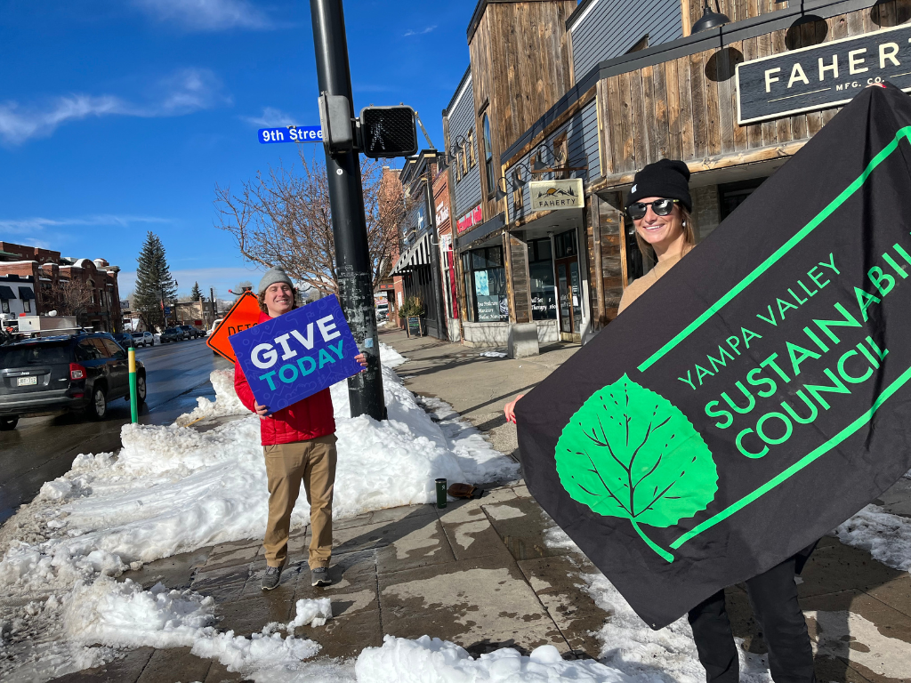 Two people stand outside holding a sign with the YVSC logo and a sign that days "Give today"