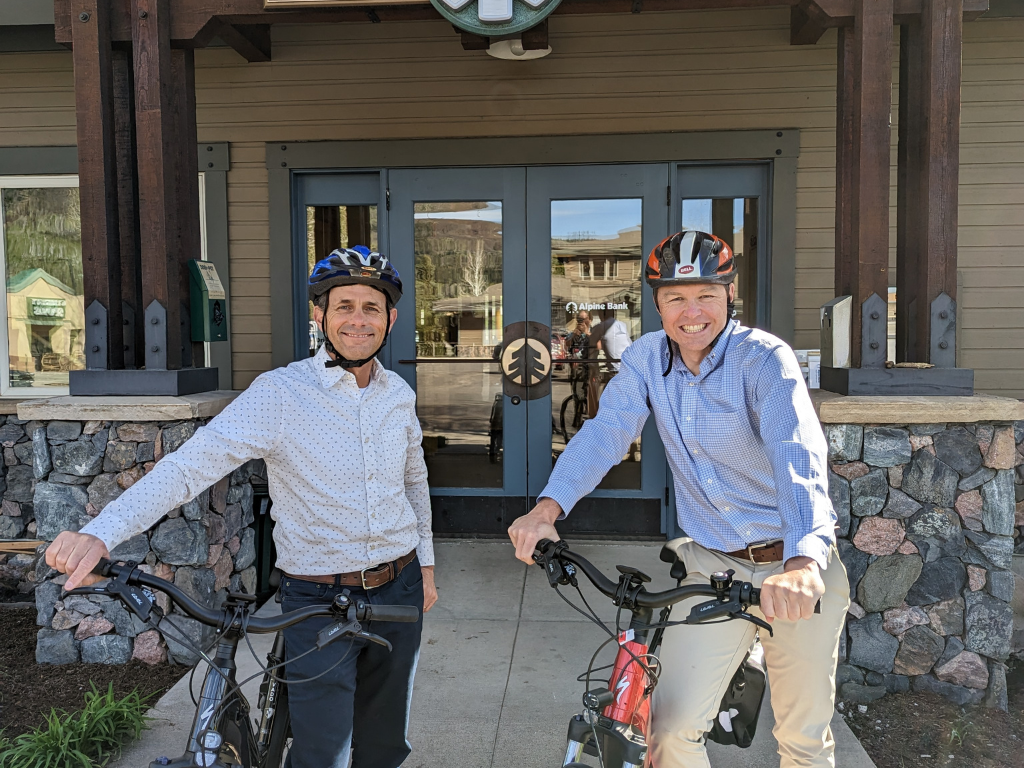 Photo of two people in suits posing in front of Alpine Bank with bikes and bike helmets.